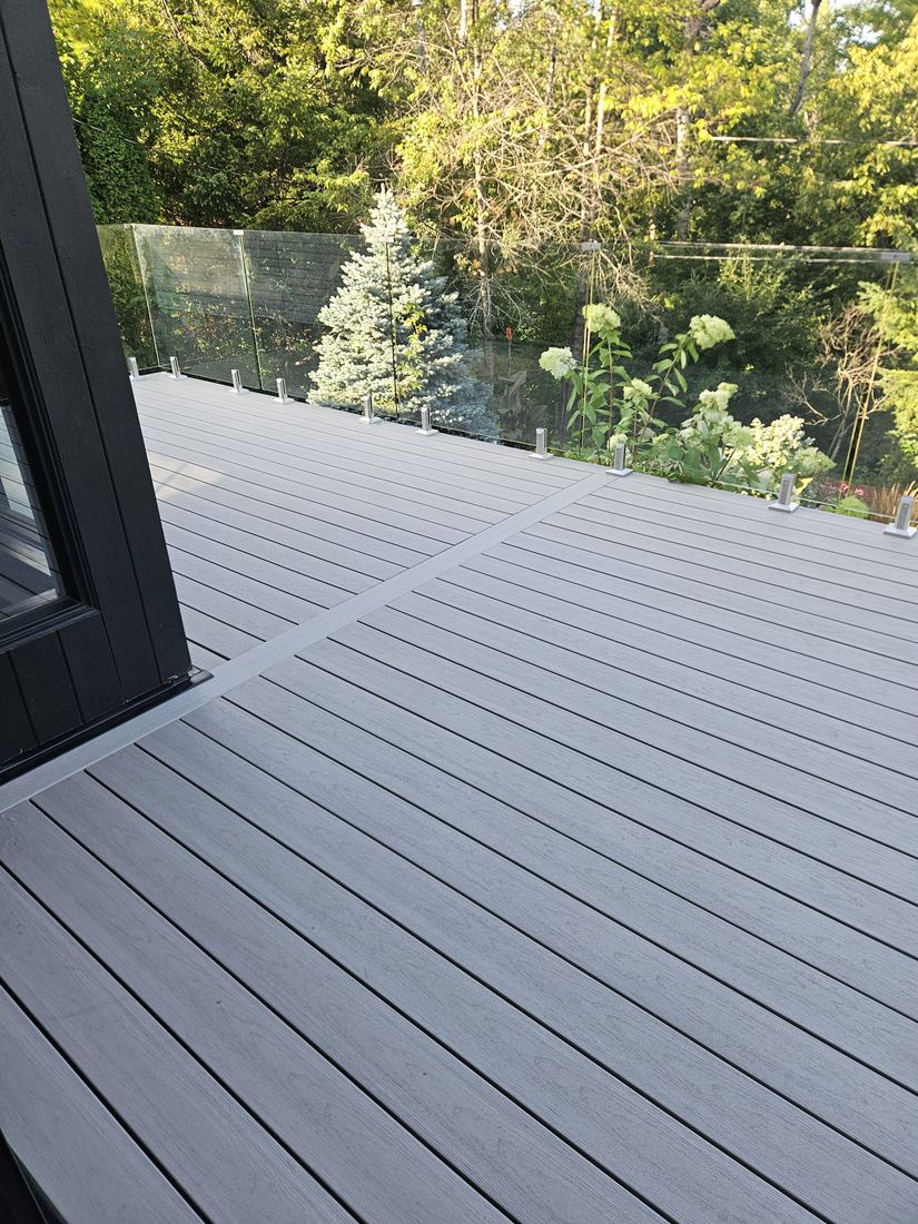 Grey composite deck with glass railing on stainless clamps beside a black-clad house