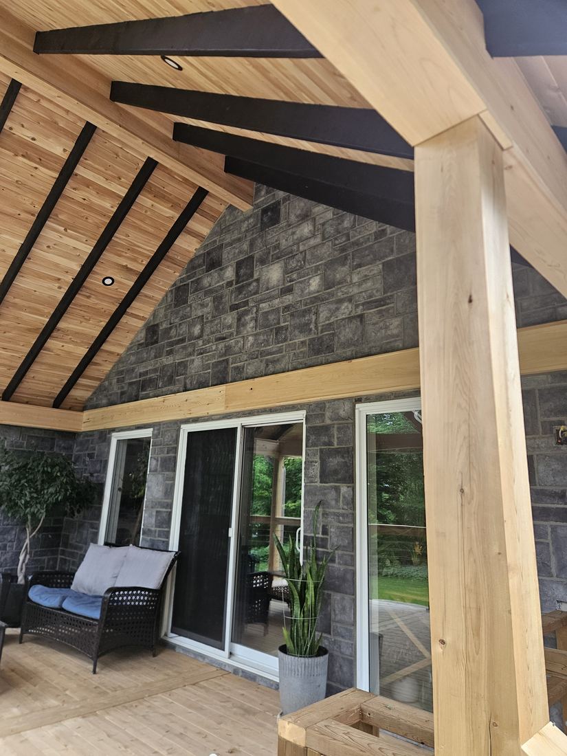 Covered deck with cedar ceiling, black beams and a wood post beside a stone wall