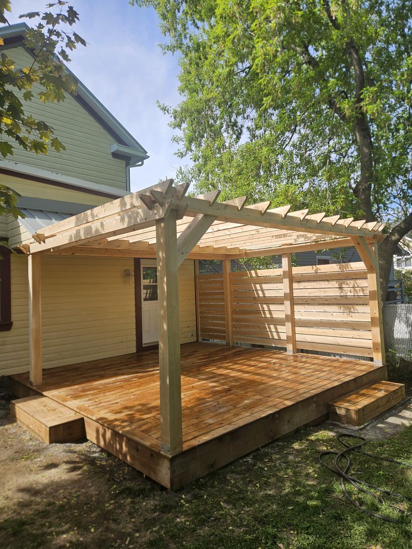 Pressure-treated wood deck with pergola and horizontal-slat privacy screen beside a yellow house