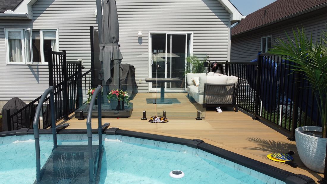 Beige composite deck beside a pool with black aluminum railing and a glass panel