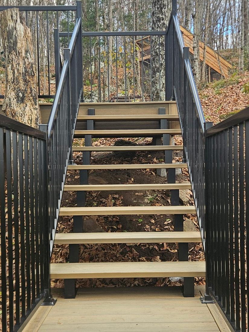 Beige composite stairs with open risers and black aluminum railings, set in a wooded area