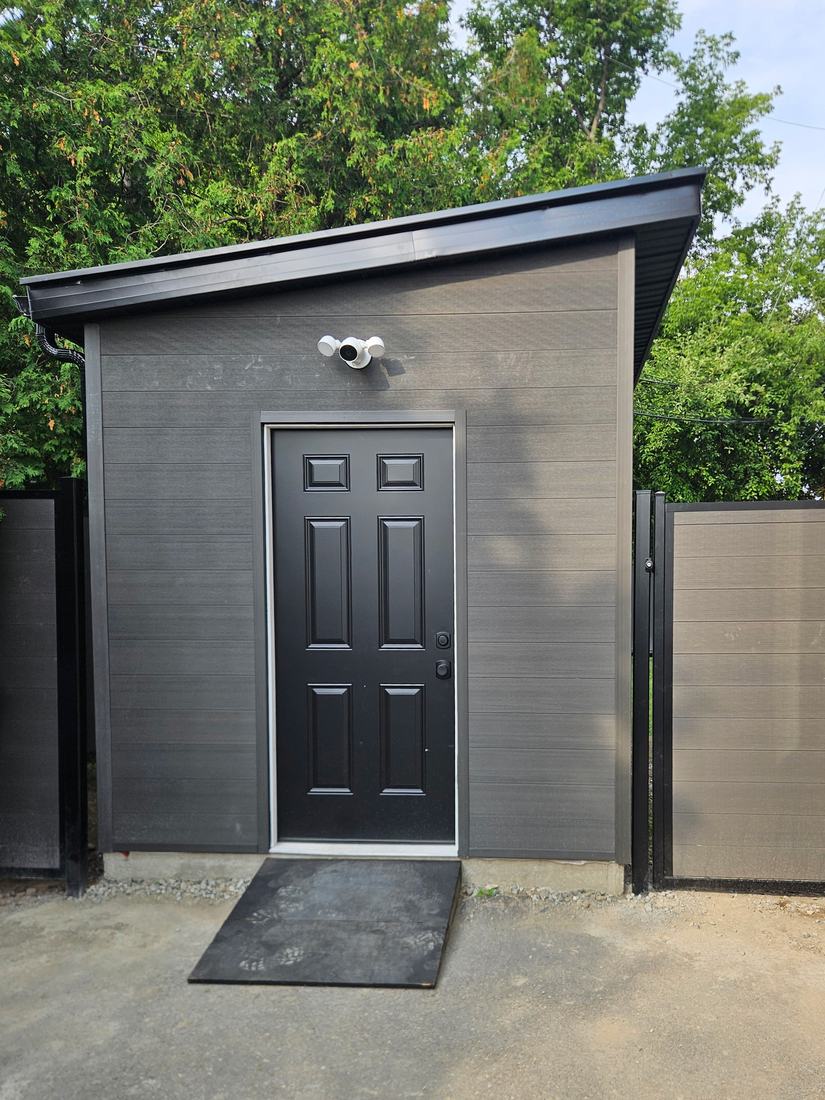 Modern shed with a sloped roof, dark grey siding, black door and a matching composite fence